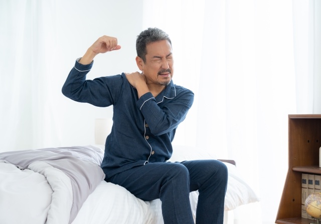 パジャマ姿でベッドに腰かけ肩を押さえて顔をしかめる男性