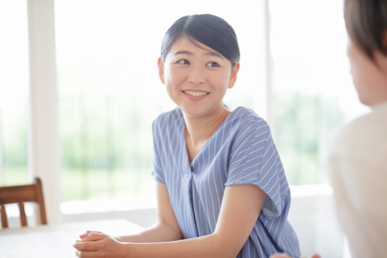 窓辺のテーブルで向かいの人と笑顔で話す青い服の女性