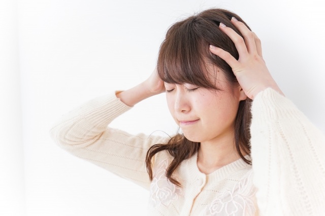 クリーム色のニットの女性が目を閉じて両手で頭を押さえる